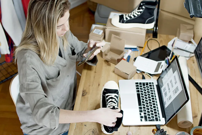 Una persona fotografía zapatillas con un teléfono junto a cajas de pedidos